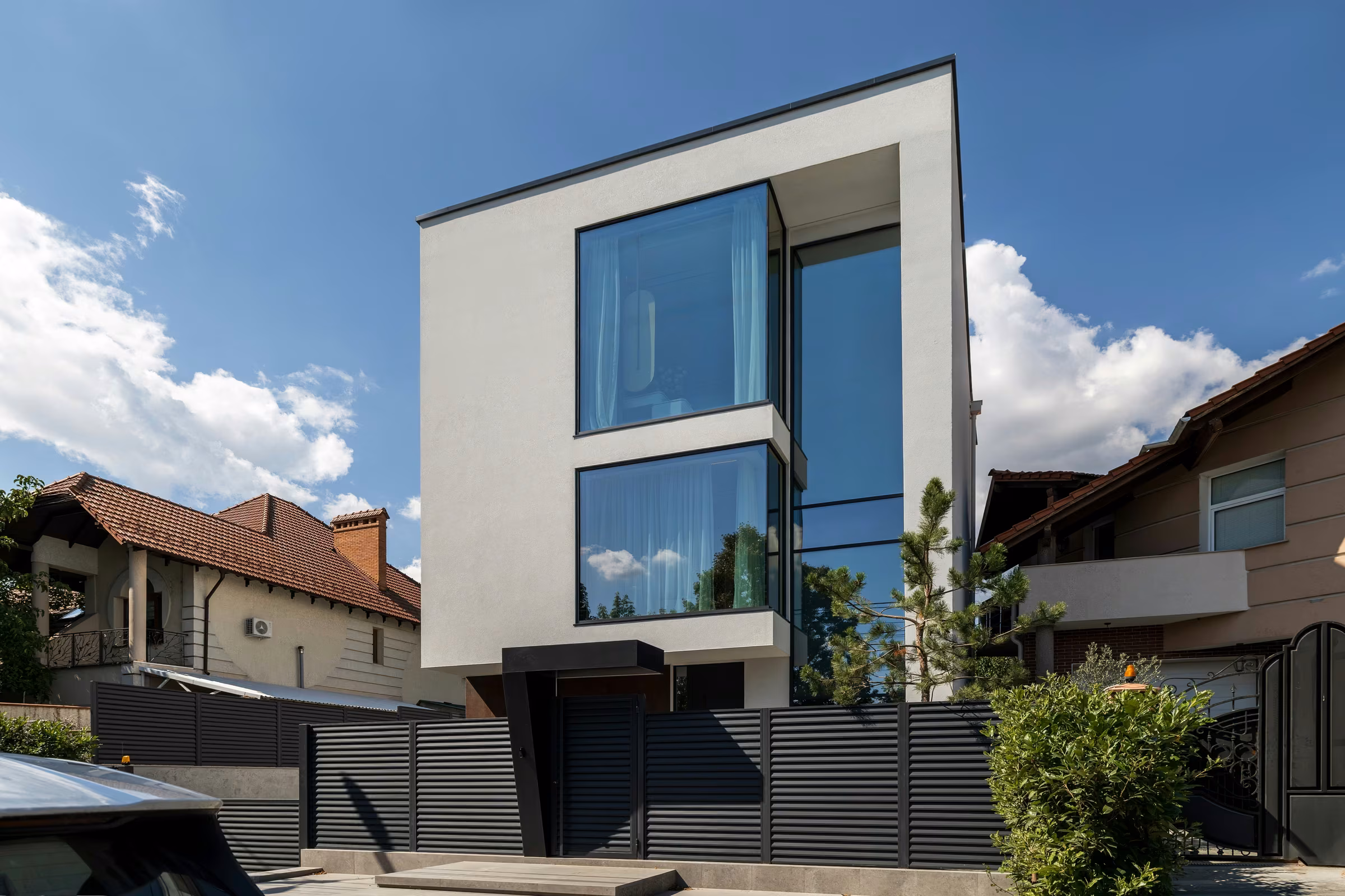 Casă contemporană cu fațadă albă, ferestre panoramice și gard metalic închis la culoare.